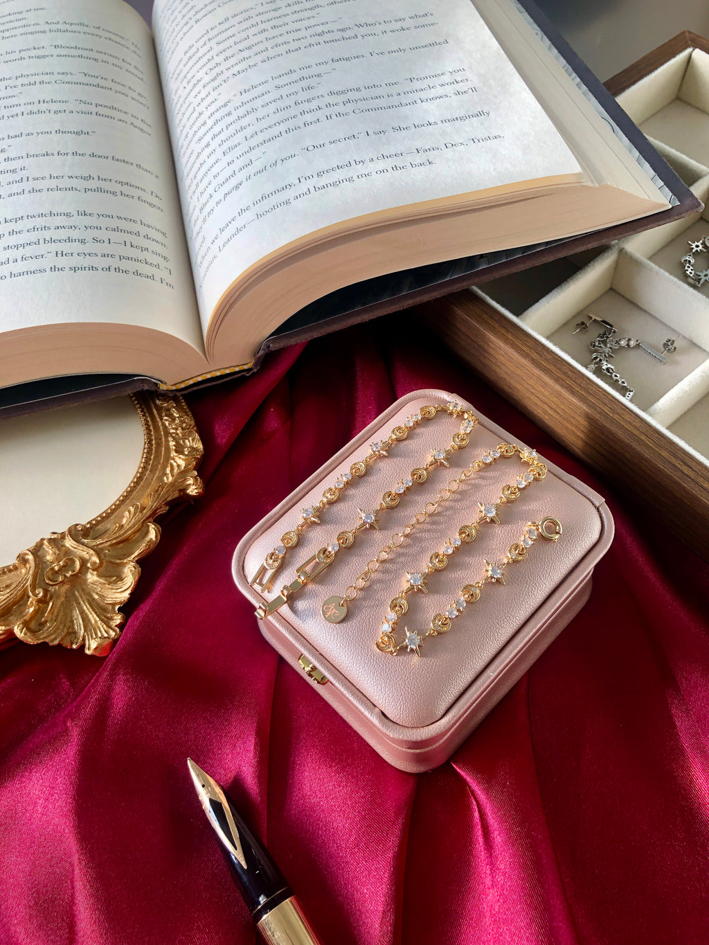 Pink jewelry box with gold bracelets on a red fabric background with an open book and fountain pen.