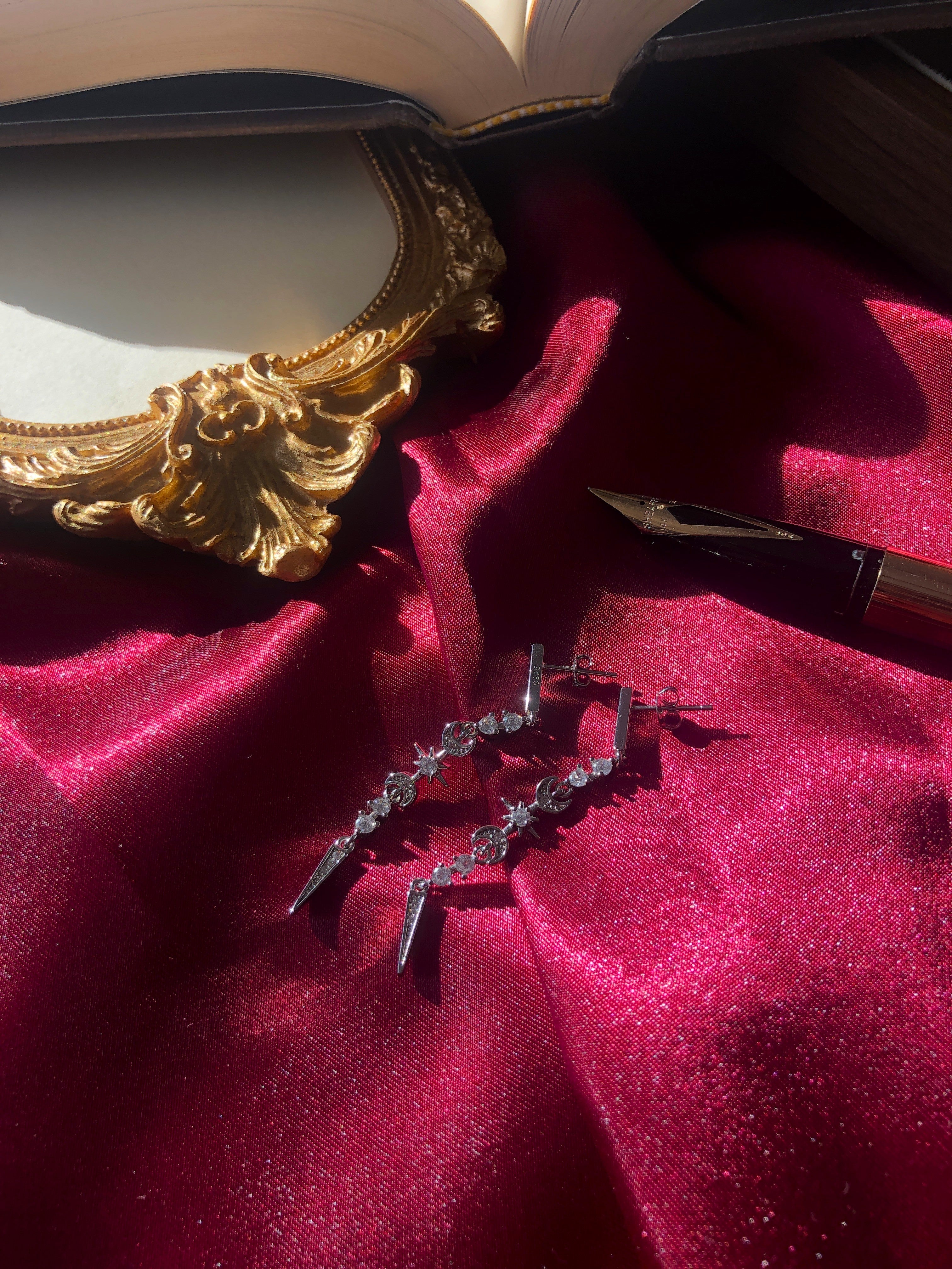 Sterling Silver Celestial ACOTAR inspired earrings on a red fabric surface with a gold frame and pen in the background